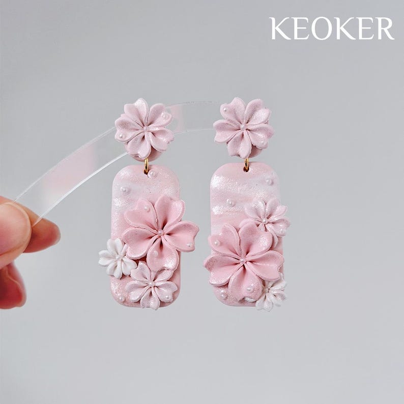 May include: Pink floral earrings with a rectangular base and gold-colored hooks. The earrings feature multiple sizes of flowers in shades of pink and white. The word "KEOKER" is printed in white in the upper right corner.