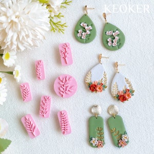 Puede incluir: Un conjunto de pendientes de arcilla polimérica con diseños florales y de hojas. Los pendientes son en tonos de verde, blanco y naranja. También hay sellos de arcilla polimérica rosa con diseños de hojas.