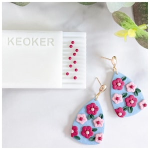 Op de afbeelding: Een paar blauwe, traanvormige oorbellen met roze en rode bloemenmotieven. De oorbellen hebben gouden haakjes.