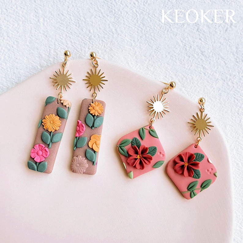 May include: A pair of gold earrings with floral designs. The earrings feature a sunburst charm and a rectangular or square shaped charm with pink, red, and green floral details.