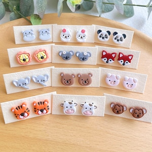 May include: A set of 10 pairs of colorful animal-shaped stud earrings. The earrings are displayed on small white cards. The animals include a hippopotamus, llama, panda, lion, koala, fox, elephant, bear, cow, and monkey.