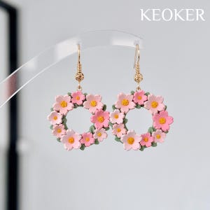 May include: Floral wreath earrings with pink flowers and yellow centers, accented with green leaves. The earrings are suspended from gold-colored hooks. The word "KEOKER" is displayed in white text.