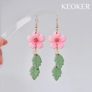 May include: A pair of pink flower earrings with gold-colored hooks and accents. Each earring features a pink flower with a gold center, a small pink bead, and two green leaves. The word "KEOKER" is in the upper right corner.