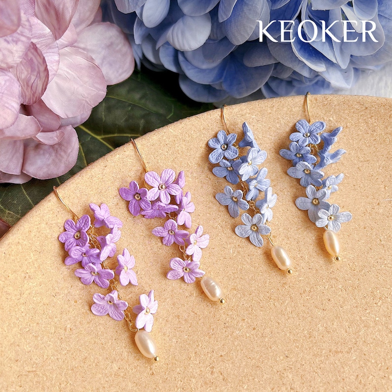 Puede incluir: Dos pares de pendientes colgantes con un racimo de peque&ntilde;as flores moradas y azules. Cada pendiente tiene una sola perla colgando de la parte inferior.
