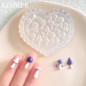 May include: A clear heart-shaped silicone mold with multiple mushroom shapes. The mold is on a white surface. Three purple and white mushroom-shaped polymer clay charms are on a hand.