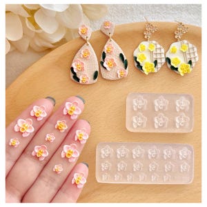 May include: A collection of handmade floral earrings and nail art decorations. The earrings feature pink and yellow flowers with green leaves. The nail art includes small, detailed flower designs in various sizes. The items are displayed on a wooden surface.