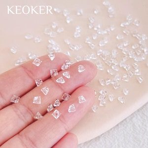 Puede incluir: Rhinestones de cristal transparente y facetado de varias formas y tamaños están dispersos sobre una superficie. Los rhinestones se sostienen en una mano, con el nombre de la marca "KEOKER" visible en la esquina superior izquierda.