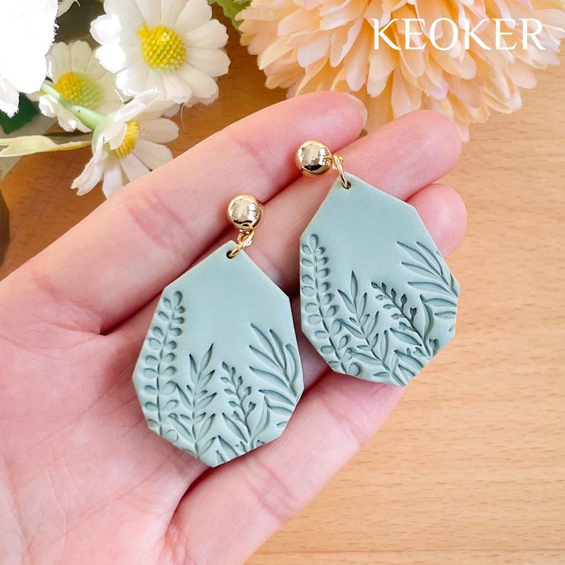Op de afbeelding: Een paar groene hangende oorbellen met een bladontwerp. De oorbellen zijn gemaakt van polymer clay en hebben goudkleurige haakjes.