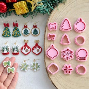 May include: A collection of Christmas-themed earrings and cookie cutters. The earrings feature designs like Christmas trees, snow globes, and ornaments. The cookie cutters are pink and shaped like bows, trees, ornaments, and snowflakes.