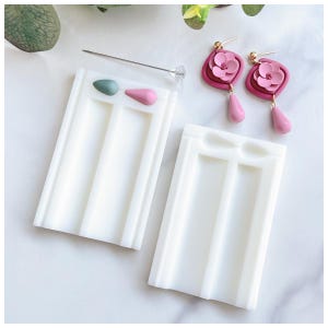 May include: Two white silicone moulds for making earrings. The moulds have two rows of teardrop-shaped cavities. There are two pink and green teardrop-shaped earrings in the top mould.