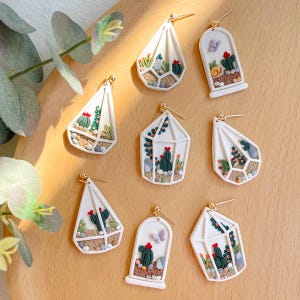 Puede incluir: Un conjunto de ocho pendientes de cerámica blanca con forma de pequeños invernaderos con diversas plantas verdes y rojas, piedras y mariposas en su interior. Los pendientes están sobre una superficie blanca.