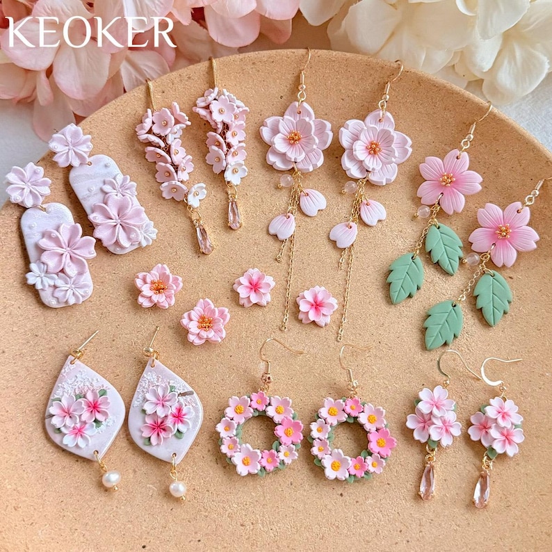 May include: A collection of handmade earrings featuring delicate pink flower designs. Styles include studs, dangles, and hoops, with accents like pearls and green leaves. The earrings are arranged on a tan surface, showcasing the variety of floral jewelry.