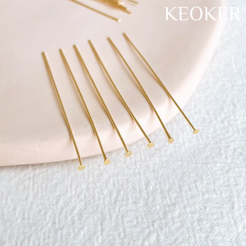 May include: A close-up of a group of gold head pins on a light pink surface. The head pins are arranged in a row, with the heads facing the viewer. The head pins are about 2 inches long.