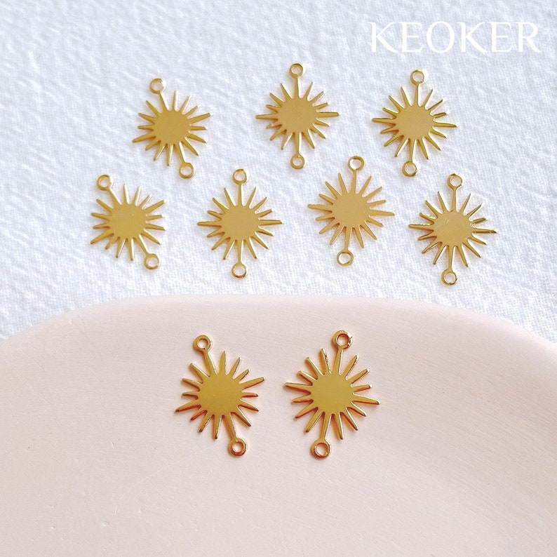 May include: Ten gold-coloured metal sunburst charms with two jump rings on each charm. The charms are arranged on a white and pink surface.