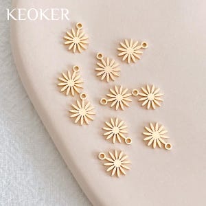 Puede incluir: Diez abalorios de metal de color dorado en forma de margarita con un bucle en la parte superior y un anillo de salto en el lateral. Los abalorios están dispuestos de forma dispersa sobre una superficie rosa pálida.