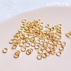 Op de afbeelding: Een verzameling kleine, goudkleurige sprongringen verspreid over een lichtroze oppervlak. De ringen zijn cirkelvormig en lijken van metaal te zijn. Het woord "KEOKER" staat in het wit rechtsboven gedrukt. Dit zijn waarschijnlijk benodigdheden voor het maken van sieraden.