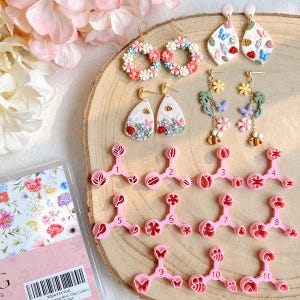 Puede incluir: Una colección de pendientes hechos a mano y cortadores de arcilla rosa. Los pendientes presentan diseños florales, de mariposas y abejas. Los cortadores de arcilla están numerados y tienen varias formas florales y de hojas.