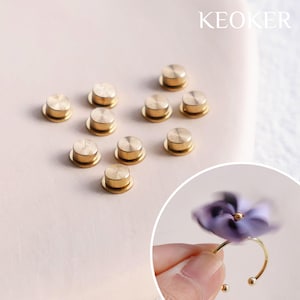 Puede incluir: Un conjunto de diez botones magnéticos de metal de color dorado. Los botones son redondos y tienen una parte superior plana. Los botones están dispuestos en círculo sobre una superficie rosa claro. La palabra "KEOKER" está impresa en negro sobre fondo blanco en la esquina superior derecha de la imagen.