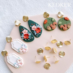 May include: Assortment of handmade polymer clay earrings featuring floral, woodland, and geometric designs. The earrings are green, white, red, and brown with gold accents. The earrings are displayed on a pink surface with gold earring backs.