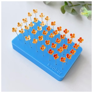 Peut inclure: Un coussin à épingles rectangulaire bleu présente de nombreuses épingles en forme de fleurs orange. Chaque épingle comporte un petit centre doré. Les épingles sont disposées en rangées soignées, créant un affichage visuellement attrayant. Le texte "keoker" est imprimé sur le côté du coussin.