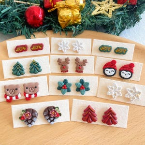 Pode incluir: Conjunto de brincos de argila polimérica com tema natalino. Os brincos apresentam vários designs, incluindo flocos de neve, renas, pinguins, árvores de Natal, azevinho e pinhas.