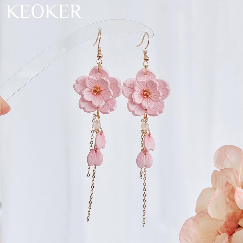May include: A pair of pink floral earrings with gold-colored accents. Each earring features a large flower with multiple petals, dangling chains, and small pink petal charms. The earrings are designed with a delicate and feminine aesthetic.