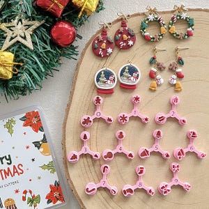 Op de afbeelding: Een verzameling kerstoorbellen en koekjessnijders. De oorbellen hebben feestelijke ontwerpen zoals ornamenten, sneeuwbollen en kransen. Roze koekjessnijders met verschillende vakantievormen worden ook weergegeven, samen met een doos met de tekst "Merry Christmas".