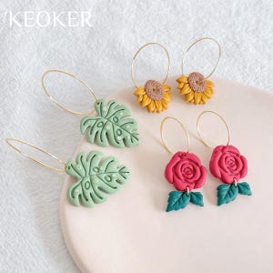 May include: A set of six gold hoop earrings with various polymer clay charms. The charms include two green monstera leaves, two red roses with green leaves, and two yellow sunflowers.
