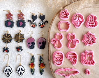 May include: A collection of Halloween-themed earrings and cookie cutters. The earrings feature designs like cauldrons, black cats, spiderwebs, skulls, and ghosts. The pink cookie cutters include shapes of ghosts, cats, bats, and skulls. The items are displayed on a wooden surface.