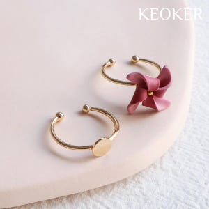 May include: Two gold rings on a pink surface. One ring has a pink flower design and the other ring has a gold disc.