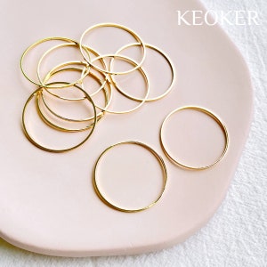 May include: A group of ten gold-colored metal hoops of varying sizes on a pink surface. The word "KEOKER" is printed in black on the surface.