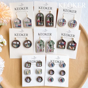 Op de afbeelding: Een verzameling polymeerklei oorbellen en oorstekers met miniatuur boekenplankontwerpen. De oorbellen tonen verschillende vormen, waaronder huizen, bogen en cirkels, met gedetailleerde boekruggen in meerdere kleuren. De oorstekers hebben geometrische vormen.