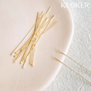 May include: A close-up of a pile of gold-colored head pins on a light pink surface. The head pins are about 2 inches long and have a small, round head.