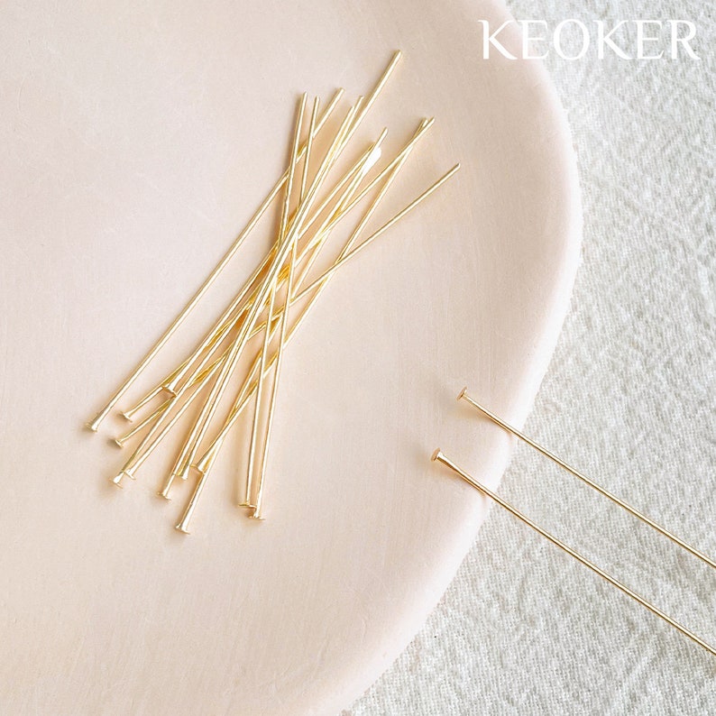 May include: A close-up of a pile of gold-colored head pins on a light pink surface. The head pins are about 2 inches long and have a small, round head.