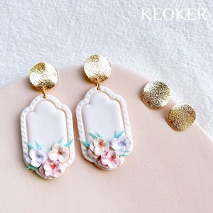 May include: A pair of white ceramic earrings with a gold accent and a floral design. The earrings are shaped like a rectangle with rounded corners and feature a cluster of pink, purple, and blue flowers.
