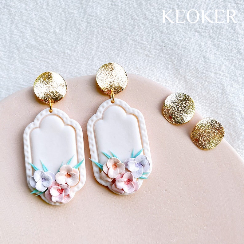 May include: A pair of white ceramic earrings with a gold accent and a floral design. The earrings are shaped like a rectangle with rounded corners and feature a cluster of pink, purple, and blue flowers.
