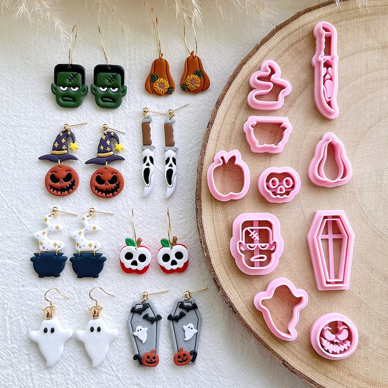 May include: A collection of Halloween-themed earrings and cookie cutters. The earrings feature designs like Frankenstein's monster, pumpkins, ghosts, and coffins. The cookie cutters are in the shapes of a knife, ghost, pumpkin, and apple. The earrings are on gold hoops.