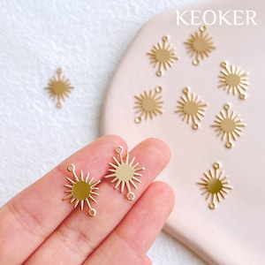 May include: Gold star-shaped charms with a hole at the top and two holes on the sides. The charms are on a pink surface and a hand is holding two of the charms.