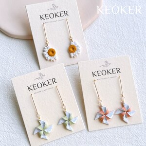 Peut inclure: Quatre paires de boucles d'oreilles en argile polym&egrave;re pr&eacute;sent&eacute;es sur des cartes. Les boucles d'oreilles pr&eacute;sentent un motif de tournesol, un motif de moulin &agrave; vent vert et blanc et un motif de moulin &agrave; vent rose et blanc. Les cartes sont &eacute;tiquet&eacute;es "KEOKER Polymer Clay Earring".