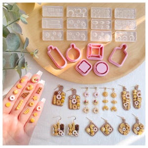 May include: A set of pink silicone moulds for making various shapes of cookies, including doughnuts, squares, and rectangles. The moulds are on a wooden surface with a variety of small, colourful cookies. There are also several pairs of earrings with cookie-shaped charms.