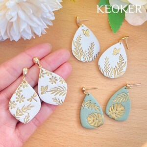 May include: Four pairs of dangle earrings with gold accents and a white or green background. The earrings feature floral and leaf designs with butterflies and dragonflies.