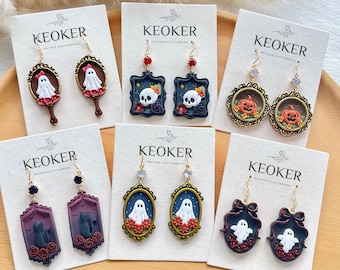 Könnte beinhalten: Sechs Paar Ohrringe aus Polymer-Ton auf KEOKER-Präsentationskarten. Die Designs umfassen Geister in Spiegeln, Totenköpfe in Rahmen, Kürbisse in verzierten Kreisen, schwarze Katzen in violetten Rahmen, Geister in ovalen Rahmen und Geister mit Schleifen.