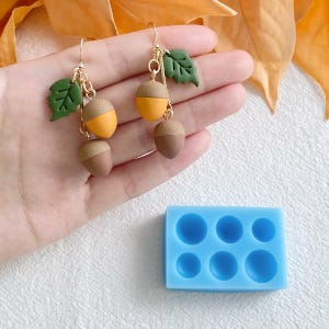 Peut inclure: Une paire de boucles d'oreilles en or avec des breloques en forme de glands et des accents de feuilles vertes. Les boucles d'oreilles sont présentées sur une main, et un moule en silicone bleu avec six cavités en forme de glands est présenté en dessous.