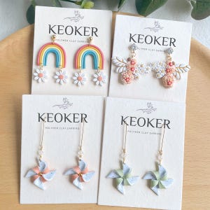 Pode incluir: Quatro pares de brincos de argila polimérica em cartões brancos com o nome da marca "KEOKER" impresso em cada cartão. Os brincos são um arco-íris com flores brancas, uma abelha com flores, uma roda de vento rosa e branca e uma roda de vento verde e branca.