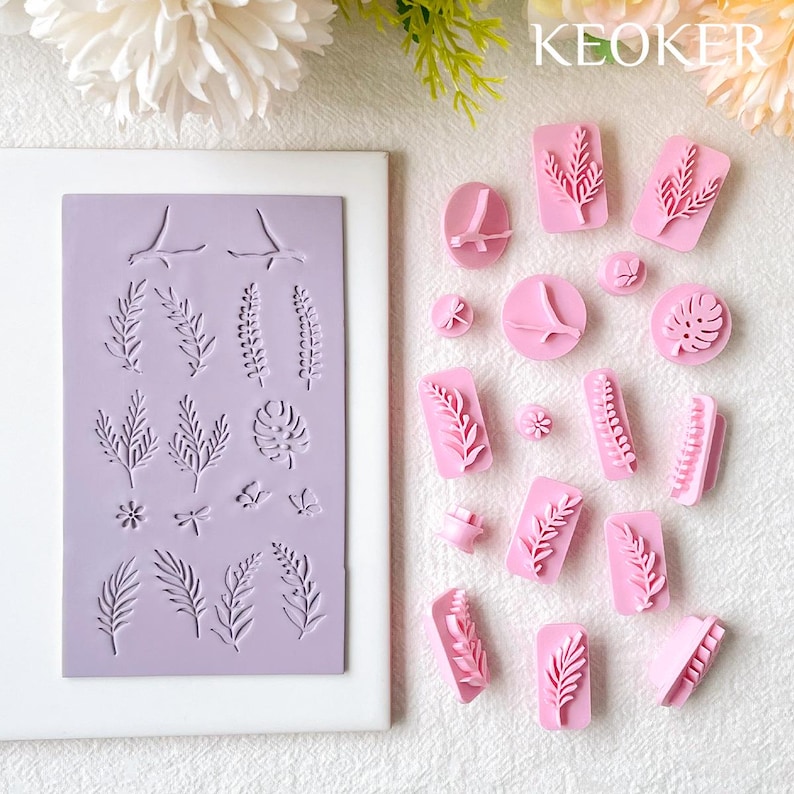 May include: A set of 20 pink silicone stamps featuring various leaf and floral designs. The stamps are arranged on a white surface with a purple silicone sheet in the background. The sheet has the same designs as the stamps. The text "KEOKER" is printed in white on a pink background in the upper right corner of the image.