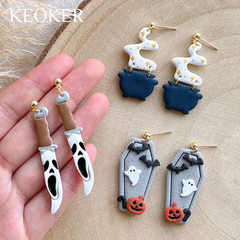 May include: Halloween-themed earrings in various designs. Includes earrings shaped like a knife with a ghost face, a cauldron with smoke, and a coffin with a ghost, bats, and a jack-o'-lantern. Gold-colored earring posts.
