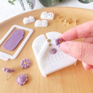Pode incluir: Uma ferramenta de pl&aacute;stico branco com um padr&atilde;o de grade e a palavra "Keoker" impressa nela. Uma m&atilde;o est&aacute; colocando um brinco em forma de flor roxo na ferramenta. H&aacute; outros brincos em forma de flor roxo e um molde de pl&aacute;stico branco na superf&iacute;cie.