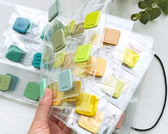 May include: A clear plastic storage binder with multiple pockets filled with small square ceramic tiles. The tiles are various shades of green, yellow, and blue. Each tile has the word "Keoker" embossed on it.