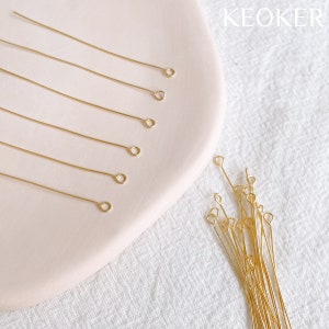 Puede incluir: Alfileres de cabeza dorada con un bucle en el extremo. Los alfileres están dispuestos sobre una superficie blanca y un plato rosa. El texto "KEOKER" es visible en la esquina superior derecha de la imagen.