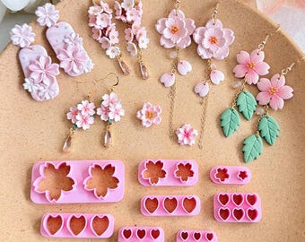 Può includere: Una collezione di orecchini floreali rosa e strumenti per la creazione di gioielli. Gli orecchini presentano vari design, tra cui grappoli di fiori, petali pendenti e foglie verdi. Sono presenti anche stampi in plastica rosa a forma di fiore e cuore.